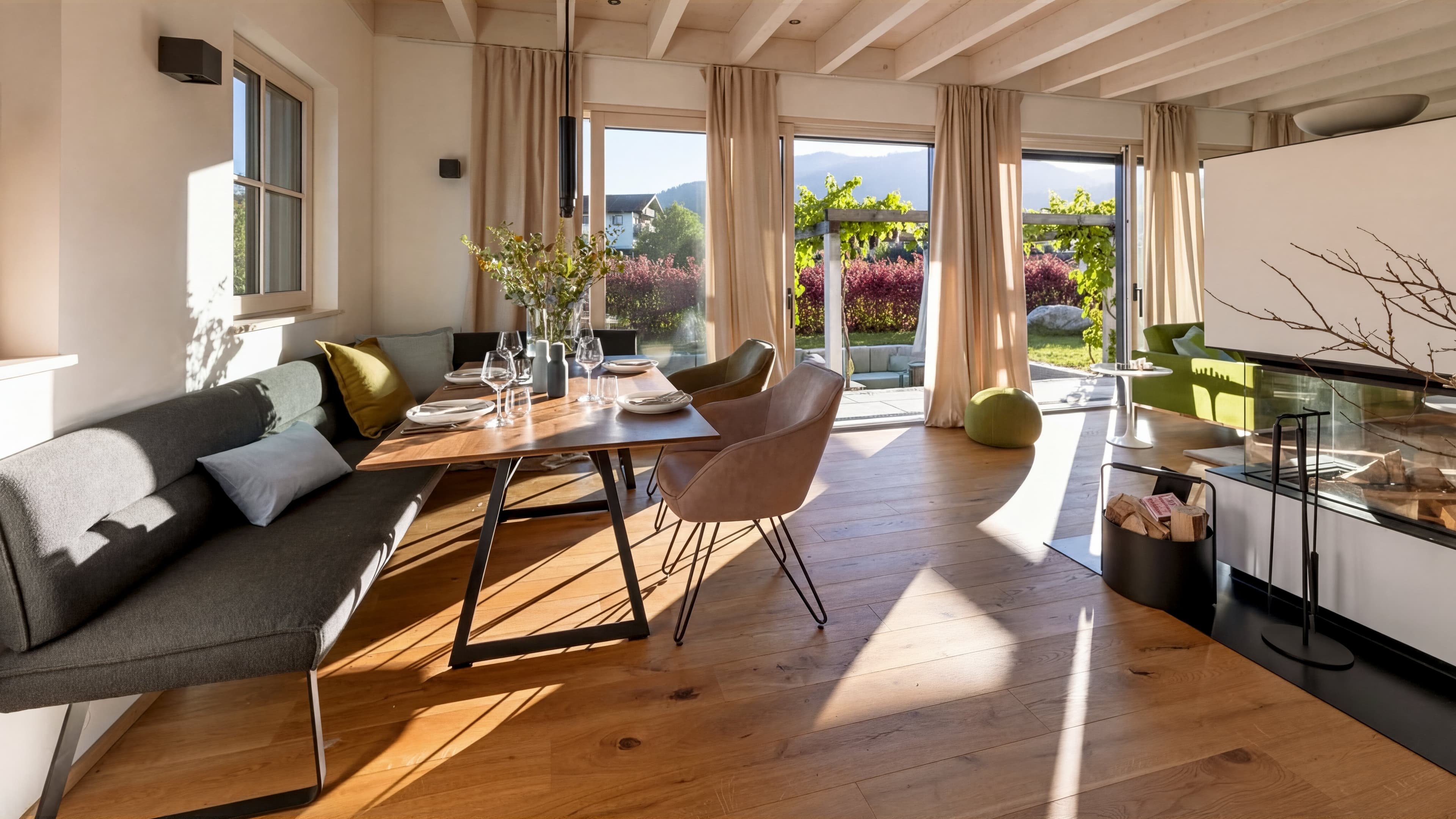 Im Essbereich am Traumhaus am Chiemsee befindet sich eine große dunkle Sitzecke mit grünen Farbakzenten an einem großen Holztisch. Gegenüber stehen zwei Stühlen in rosa und dunkelgrau. Bodentiefe Fenster erhellen den Raum und Sonnenlicht strahlt ein.