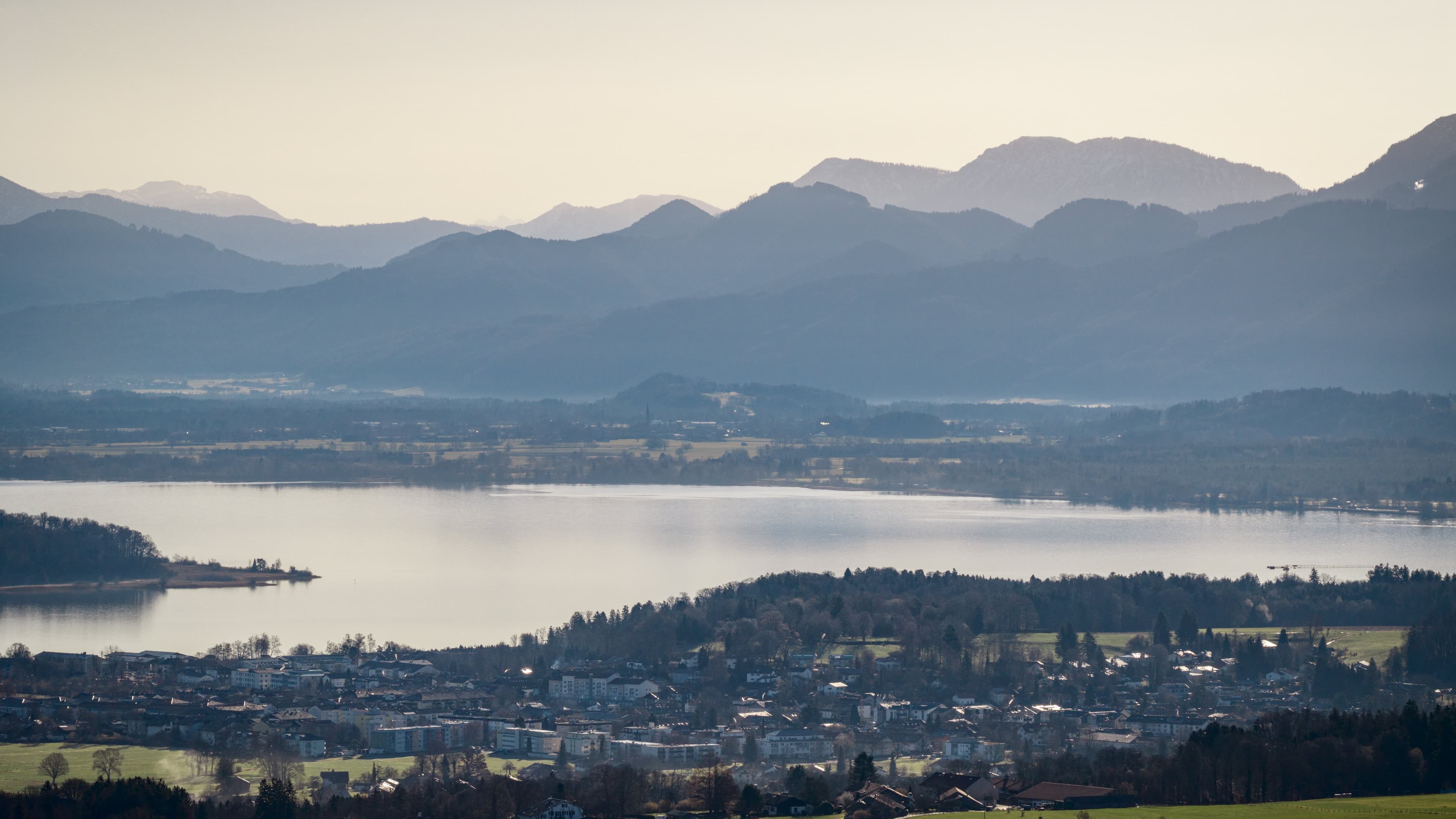 Blick von einem Aussichtspunkt am Chiemsee. Im Vordergrund befindet sich ein kleines bayerisches Örtchen, dahinter erstreckt sich der Chiemsee und die majestätische Alpenlandschaft bei sommerlichen Wetter.
